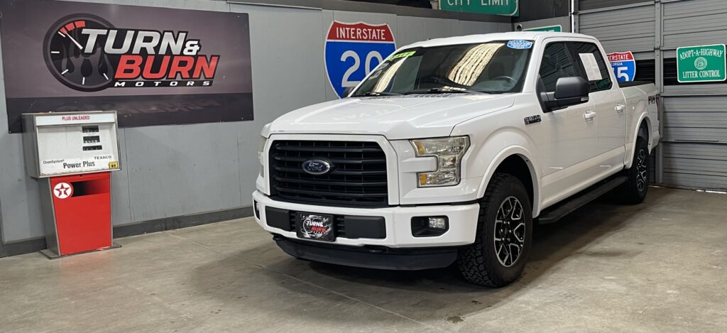 White Ford F-150 pickup on showroom floor beside dealership signage and a gas-pump display, with Turn & Burn Motors banner in the background.