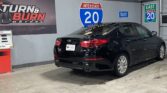 Black Kia Optima parked indoors beside a Turn & Burn Motors banner and large I-20 Interstate signs.