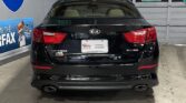 Rear view of a black Kia Optima in a dealership showroom, with red taillights and dual exhausts; wall signs (Interstate/Highway) and a blue banner to the left.